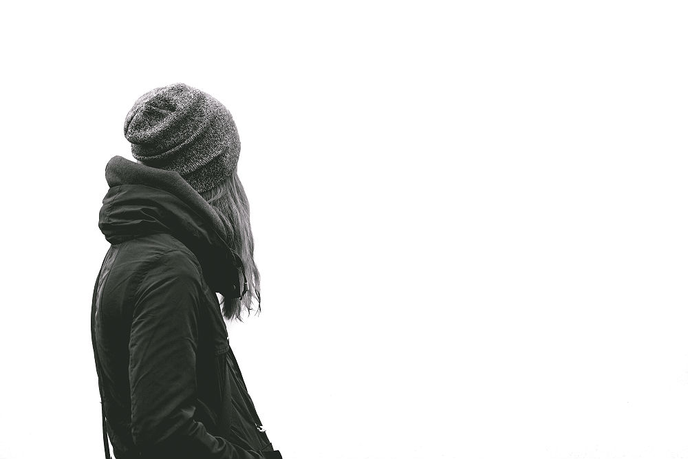 Black and white photo of a girl in winter clothes (jacket and beanie) looking away from the camera. The background is white, her hair shields her face from the view (it looks blonde), and her hands are in her pockets.
