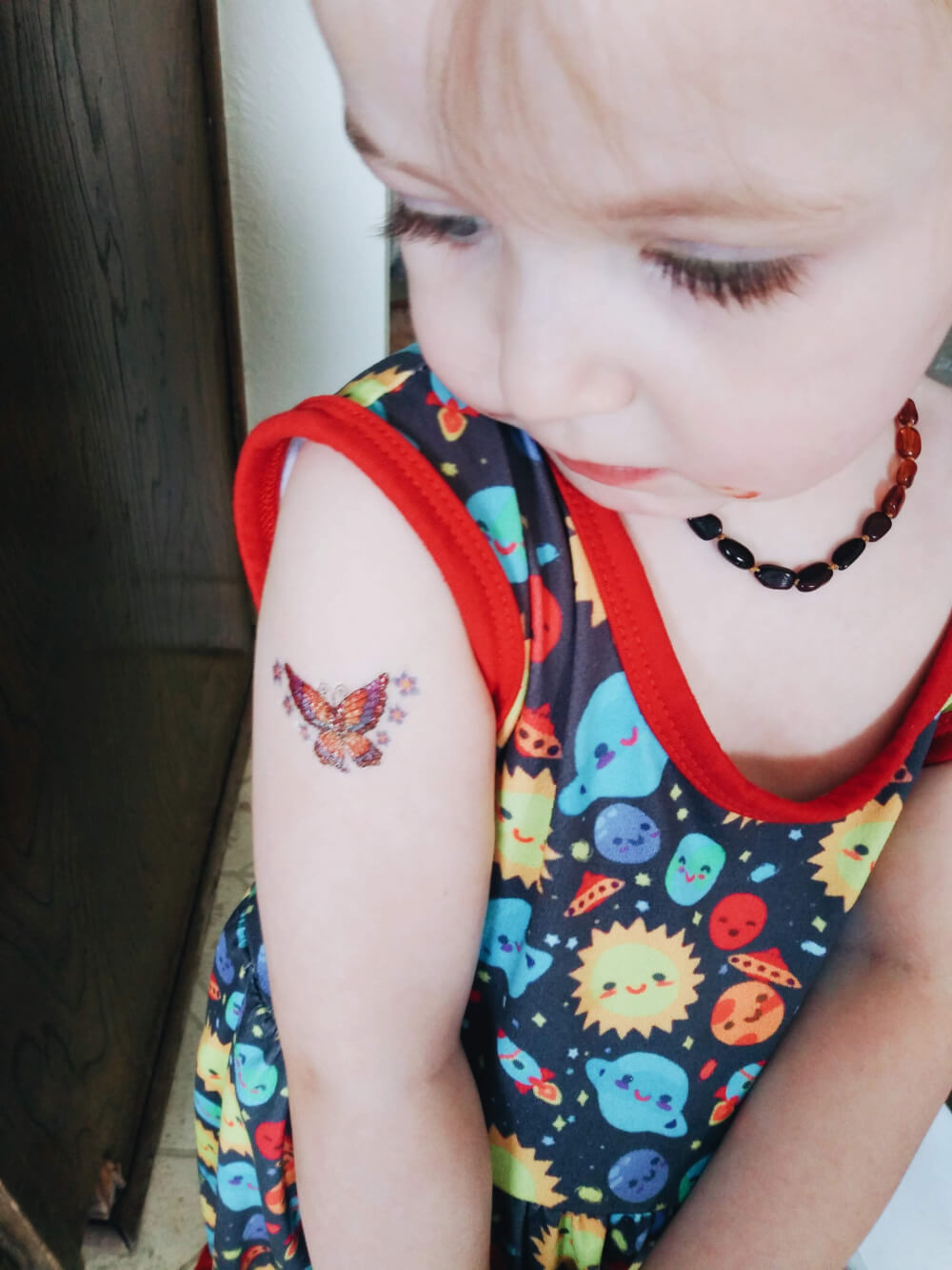 White toddler in solar system dress, looking at a temporary butterfly tattoo on her shoulder